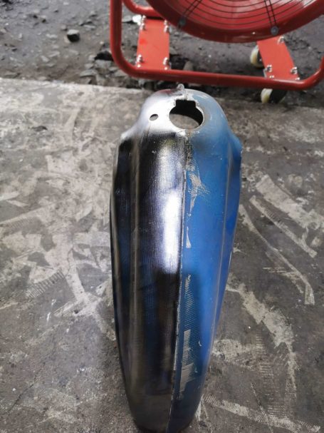 Blauer, beschädigter Motorradtank mit einer Delle und einem Loch an der Oberseite.
